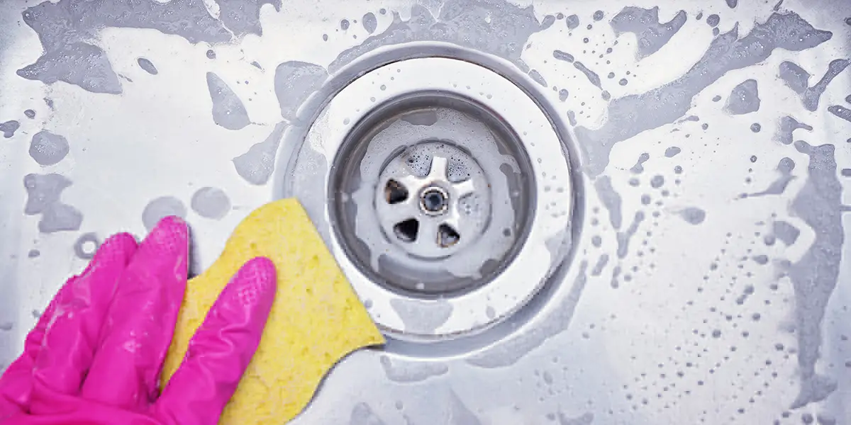 چگونه سینک آشپزخانه را تمیز کنیم,عکس بزرگنمایی شده از کاسه سینک با آب صابون و دستکش صورتی در حال تمیز کردن لگن